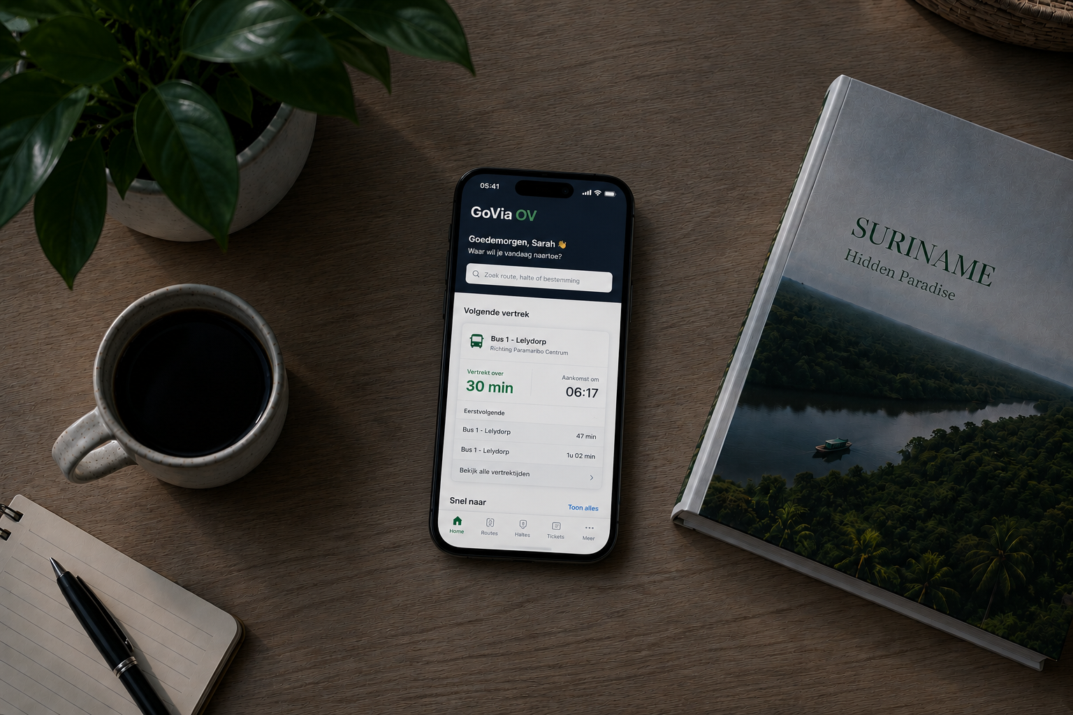 GoVia OV mobile app showing live next departure on a phone resting on a wooden table next to a coffee cup and a Suriname travel book.